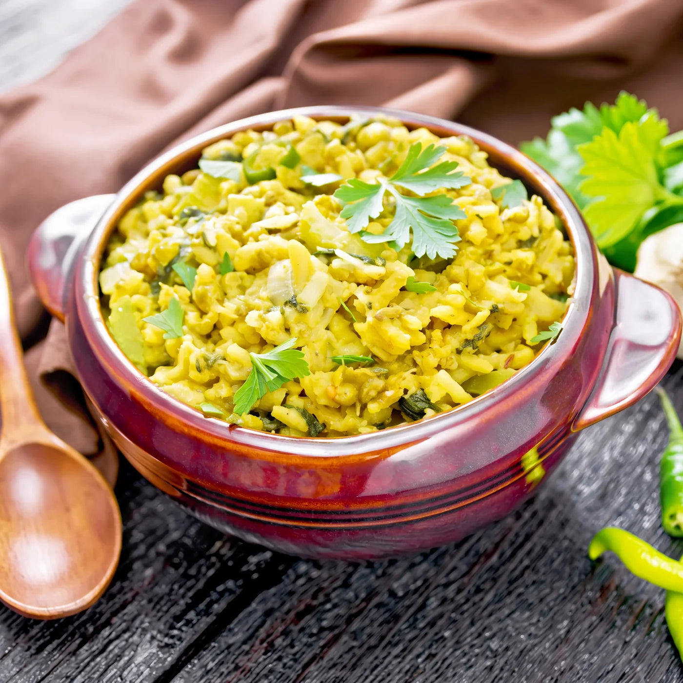 A bowl of golden kitchari topped with fresh herbs, with a wooden spoon, green chillies and garlic alongside.